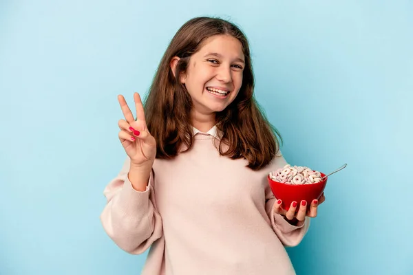 小女孩拿着一碗谷物 蓝色背景隔离 快乐而无忧无虑 用手指展示和平的象征 — 图库照片