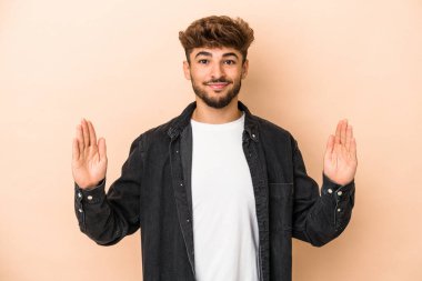 Young arab man isolated on beige background holding something little with forefingers, smiling and confident.