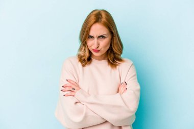 Mavi arka planda izole edilmiş genç beyaz kadın hoşnutsuzca kaşlarını çatmış, kollarını kavuşturmuş..