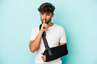 Young arab man with broken hand isolated on blue background keeping a secret or asking for silence.