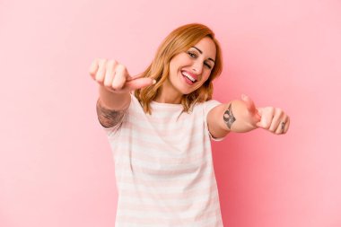 Pembe arka planda izole edilmiş genç beyaz kadın iki parmağını da kaldırıyor, gülümsüyor ve kendinden emin..