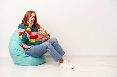 Young caucasian woman sitting on puff with popcorns isolated on white background is saying a secret hot braking news and looking aside