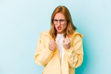 Mavi arka planda izole edilmiş genç beyaz kadın yumruğunu kameraya gösteriyor, agresif yüz ifadesi..