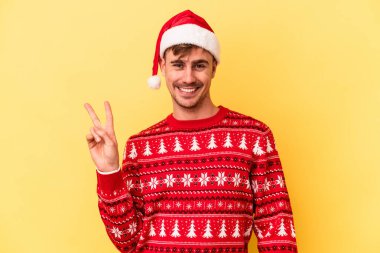 Kafkasyalı genç adam, sarı arka planda izole edilmiş bir şekilde Noel 'i kutluyor neşeli ve kaygısız parmaklarıyla barış sembolü gösteriyor..