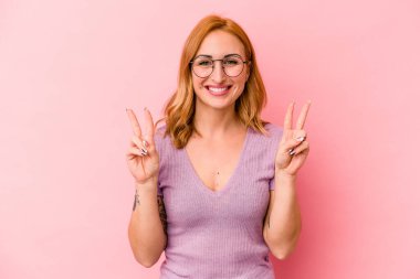 Pembe arka planda izole edilmiş genç beyaz kadın zafer işareti gösteriyor ve gülümsüyor..