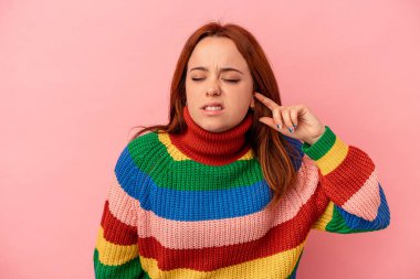 Pembe arka planda izole edilmiş genç beyaz bir kadın parmaklarıyla kulaklarını kapatıyor, gürültülü bir ortam yüzünden stresli ve çaresiz..