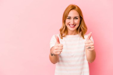 Pembe arka planda izole edilmiş genç beyaz kadın gülümsüyor ve başparmağını kaldırıyor.