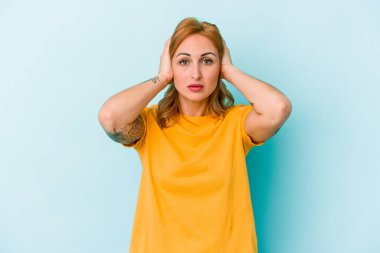 Kafkasyalı genç bir kadın mavi arka planda izole edilmiş kulakları kulaklarıyla kapatıyor çok fazla ses duymamaya çalışıyor..