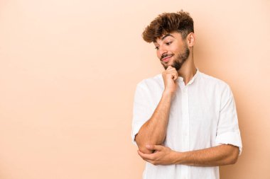 Genç Arap adam bej arka planda izole edilmiş. Şüpheli ve şüpheci bir ifadeyle yana bakıyor..