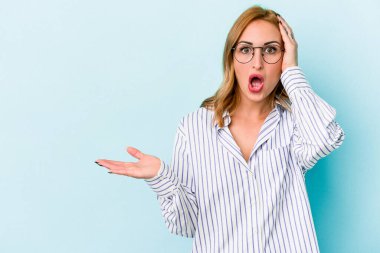 Mavi arka planda izole edilmiş genç beyaz kadın, avuç içinin fotokopi alanını tutmaktan etkilenmiş..