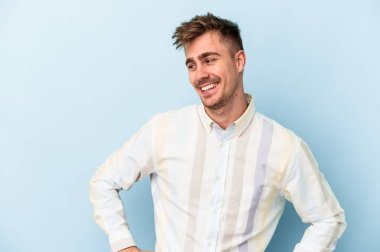 Mavi arka planda izole edilmiş genç beyaz adam mutlu bir şekilde gülüyor ve ellerini karnının üstünde tutarak eğleniyor..