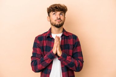 Young arab man isolated on beige background holding hands in pray near mouth, feels confident.