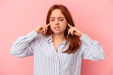Pembe arka planda izole edilmiş genç beyaz bir kadın parmaklarıyla kulaklarını kapatıyor, gürültülü bir ortam yüzünden stresli ve çaresiz..