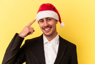 Yeni yılı kutlayan beyaz bir genç adam sarı arka planda izole edilmiş bir Noel şapkası takıyor.