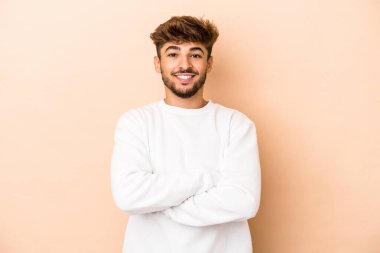 Young arab man isolated on beige background who feels confident, crossing arms with determination.