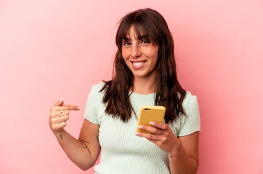 Arjantinli genç bir kadın pembe arka planda izole edilmiş bir cep telefonu tutuyor tişört fotokopi alanını işaret ediyor, gururlu ve kendinden emin.