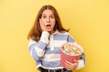 Elinde patlamış mısır tutan küçük beyaz kız sarı arka planda izole edilmiş, şaşırmış ve şok olmuş..