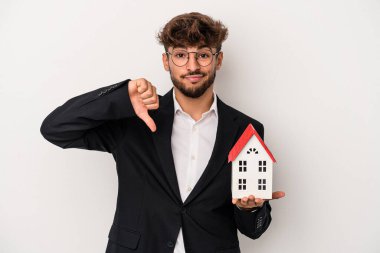 Young arab real estate man holding a model house isolated on isolated background showing a dislike gesture, thumbs down. Disagreement concept.
