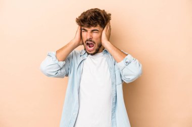 Young arab man isolated on beige background covering ears with hands trying not to hear too loud sound.