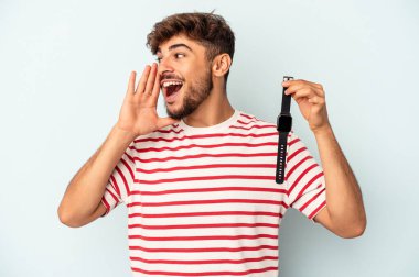 Young mixed race man holding a wrist watch isolated on blue background shouting and holding palm near opened mouth.
