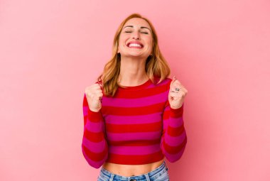 Pembe arka planda izole edilmiş genç beyaz kadın zaferi, tutkuyu ve coşkuyu kutluyor, mutlu bir ifade..