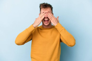 Mavi arka planda soyutlanmış genç beyaz bir adam elleriyle gözlerini kapatıyor, geniş bir gülümsemeyle bir sürpriz bekliyor..