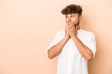 Young arab man isolated on beige background thoughtful looking to a copy space covering mouth with hand.