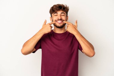 Genç melez adam gri arka planda izole edilmiş gülümsüyor, parmaklarını ağıza doğrultuyor..