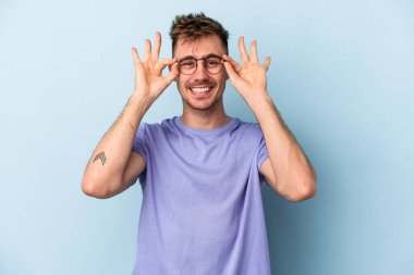 Mavi arka planda izole edilmiş genç beyaz adam gözlerinde sorun olmadığını gösteriyor.