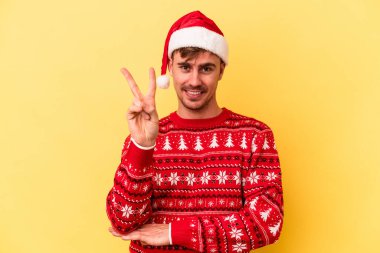 Genç beyaz adam Noel 'i kutluyor. Sarı arka planda izole edilmiş. İki numarayı parmaklarıyla gösteriyor..