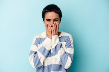 Mavi arka planda izole edilmiş genç beyaz kadın bir şeye gülüyor, elleriyle ağzını kapatıyor..