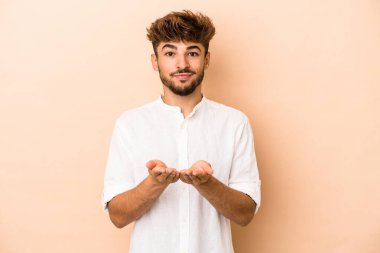 Young arab man isolated on beige background holding something with palms, offering to camera.