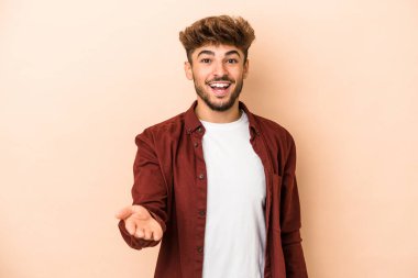 Young arab man isolated on beige background stretching hand at camera in greeting gesture.