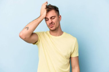 Kafkasyalı genç adam mavi arka planda izole edilmiş bir şekilde bir şeyi unutuyor. Alnına avuç içi ve gözlerini kapayarak vuruyor..