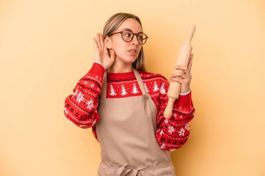 Genç fırıncı kadın Noel için kurabiye yapıyor sarı arka planda izole edilmiş bir şekilde bir dedikoduyu dinlemeye çalışıyor..