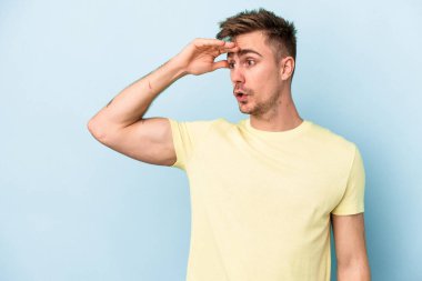 Kafkasyalı genç adam mavi arka planda izole edilmiş, uzaklara bakıyor ve elini alnına koyuyor..