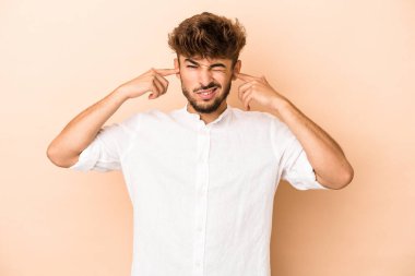 Young arab man isolated on beige background covering ears with fingers, stressed and desperate by a loudly ambient.