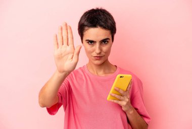 Elinde cep telefonu olan beyaz bir kadın pembe arka planda izole halde duruyor. Elini uzatmış, dur işareti veriyor ve seni engelliyor..