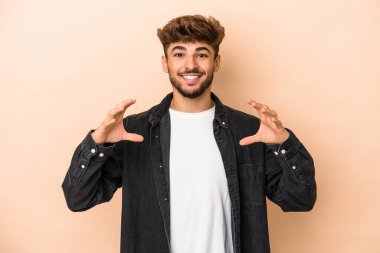 Young arab man isolated on beige background holding something with palms, offering to camera.
