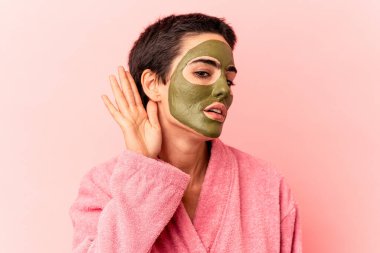 Pembe arka planda soyutlanmış yüz maskesi takan beyaz bir kadın bir dedikoduyu dinlemeye çalışıyor..