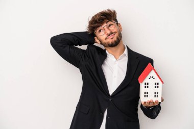 Young arab real estate man holding a model house isolated on isolated background touching back of head, thinking and making a choice.