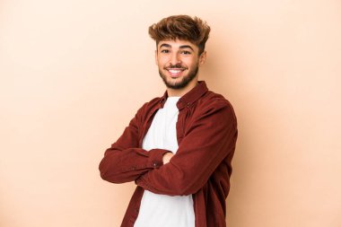 Young arab man isolated on beige background who feels confident, crossing arms with determination.