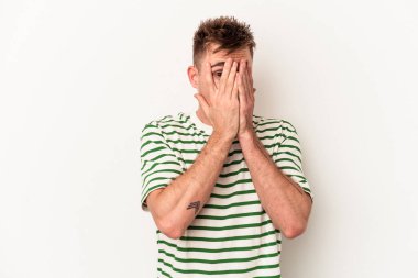 Beyaz arka planda izole edilmiş genç beyaz bir adam parmaklarının arasından korkmuş ve gergin bir şekilde göz kırpıyor..