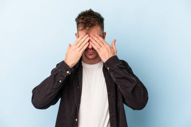 Mavi arka planda izole edilmiş genç beyaz adam elleriyle gözlerini kapatmaktan korkuyor..