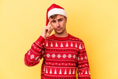 Genç beyaz adam, sarı arka planda izole edilmiş bir şekilde Noel 'i kutluyor. Şakağı işaret ediyor, düşünüyor ve bir göreve odaklanıyor..