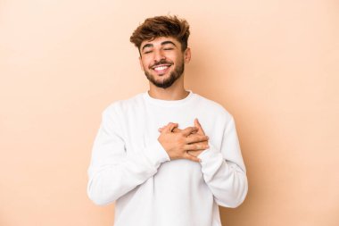 Genç Arap adam bej arka planda izole edilmiş. Gülüyor, elini kalbe koyuyor, mutluluk kavramını koruyor..