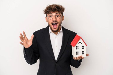 Young arab real estate man holding a model house isolated on isolated background receiving a pleasant surprise, excited and raising hands.