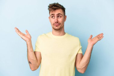 Kafkasyalı genç adam mavi arka planda izole edilmiş sorgulama jestinde şüphe ediyor ve omuz silkiyor..
