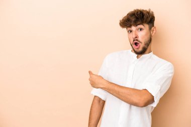 Young arab man isolated on beige background smiling and pointing aside, showing something at blank space.