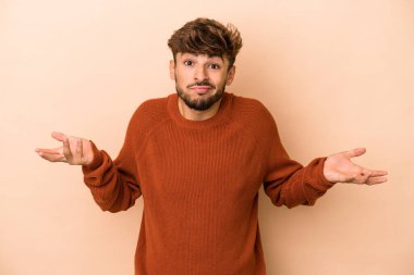 Young arab man isolated on beige background doubting and shrugging shoulders in questioning gesture.